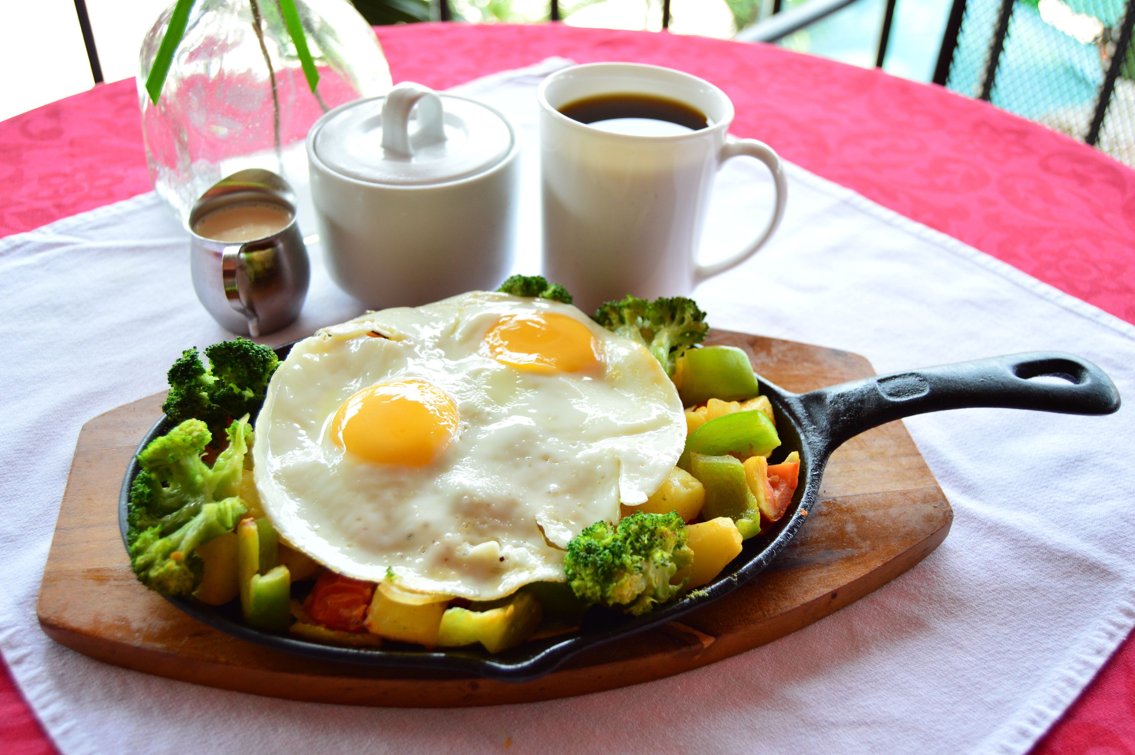 A breakfast plate with eggs, broccoli, and potatoes on a wooden board, accompanied by two cups of coffee and a small container of condiments.