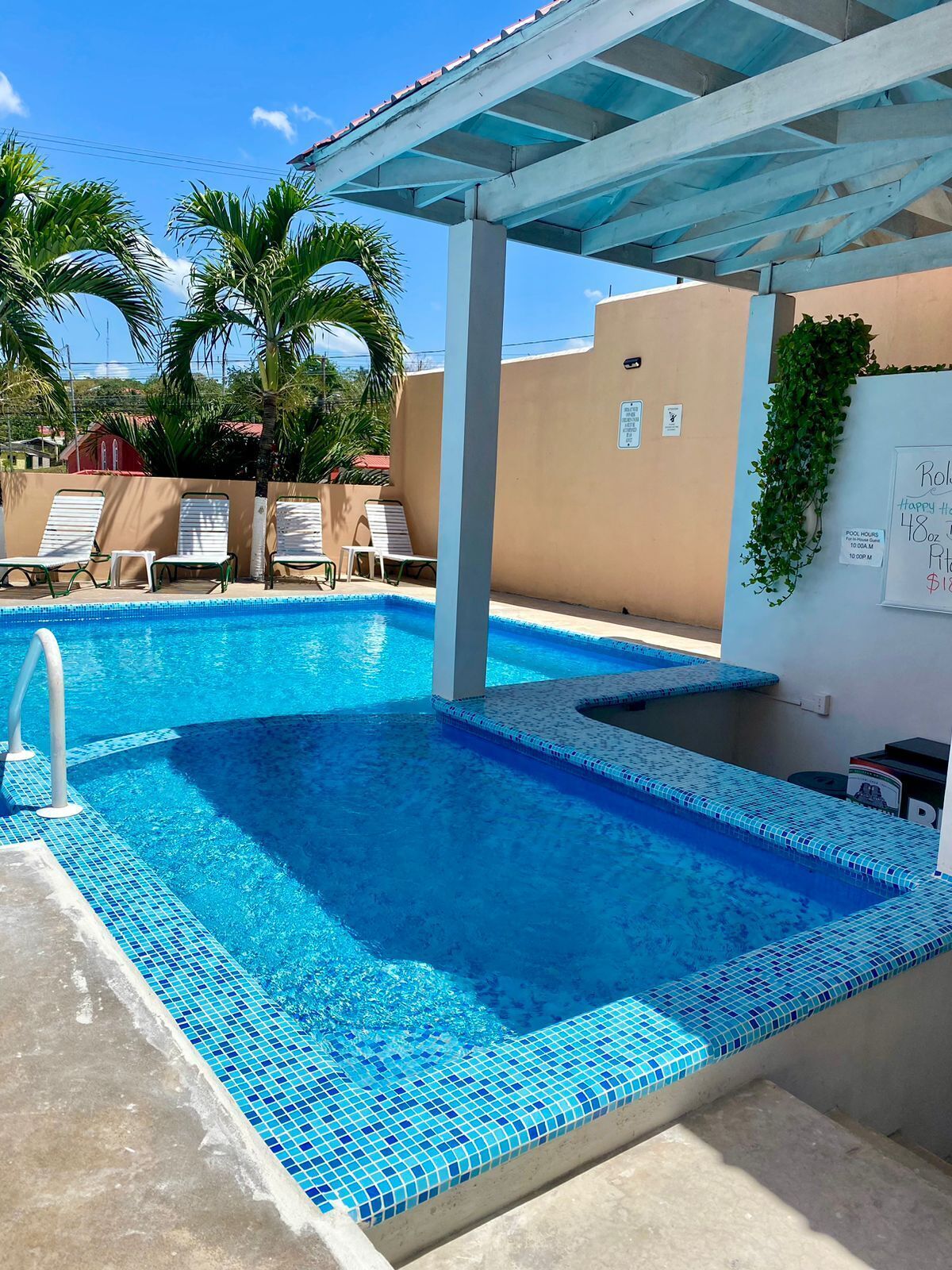 Piscina con borde de azulejos, sillas de descanso y una zona techada con un cartel que indica los horarios y tarifas.