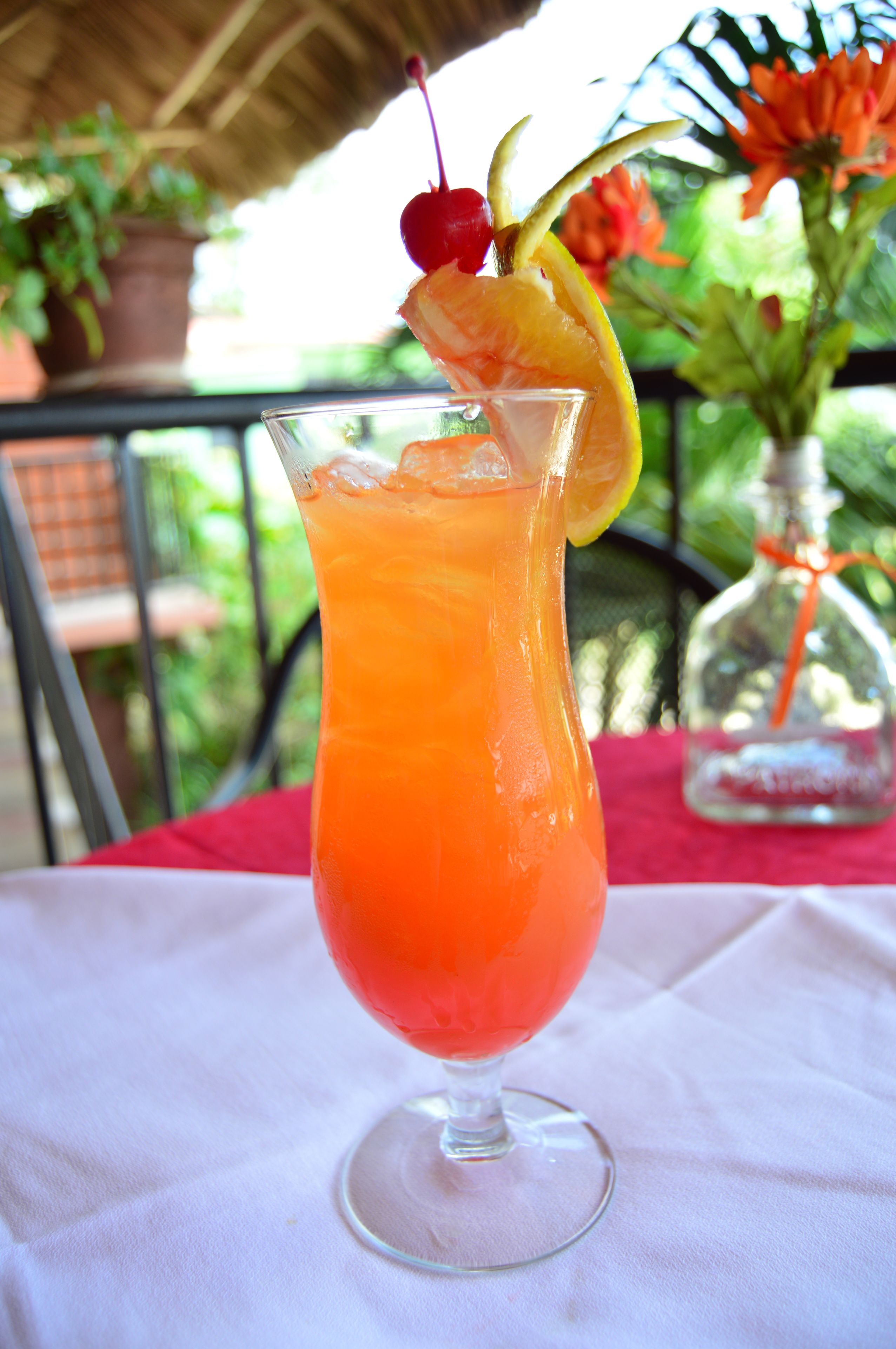 A tall glass with a red drink, garnished with a cherry and an orange slice, on a white tablecloth.