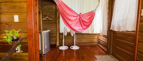 A wooden cabin interior with a hammock, white curtains, a small fan, and a plant.
