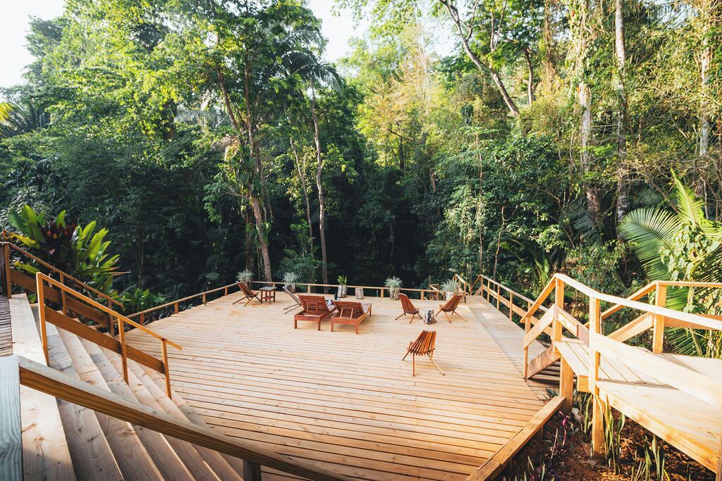 A wooden deck with outdoor seating and a railing, surrounded by dense forest.