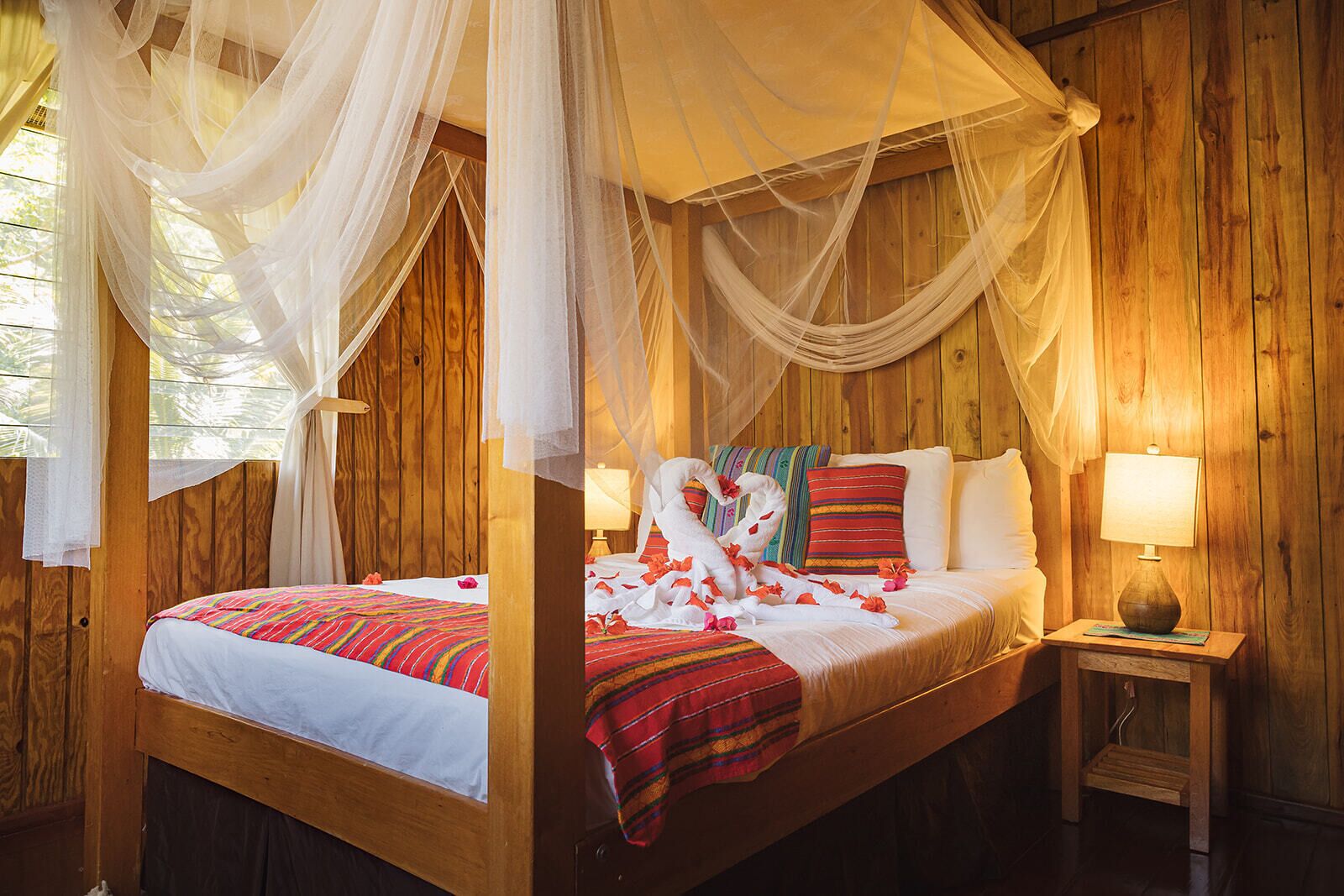 A four-poster canopy bed with white linens, a colorful striped bedspread, and a red and gold throw blanket. There is a wooden nightstand with a lamp and a small shelf.