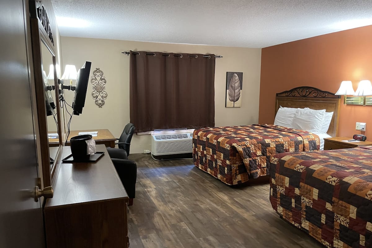 Quarto de hotel com duas camas, uma escrivaninha, uma cadeira, um ar condicionado e uma luminária fixada na parede.