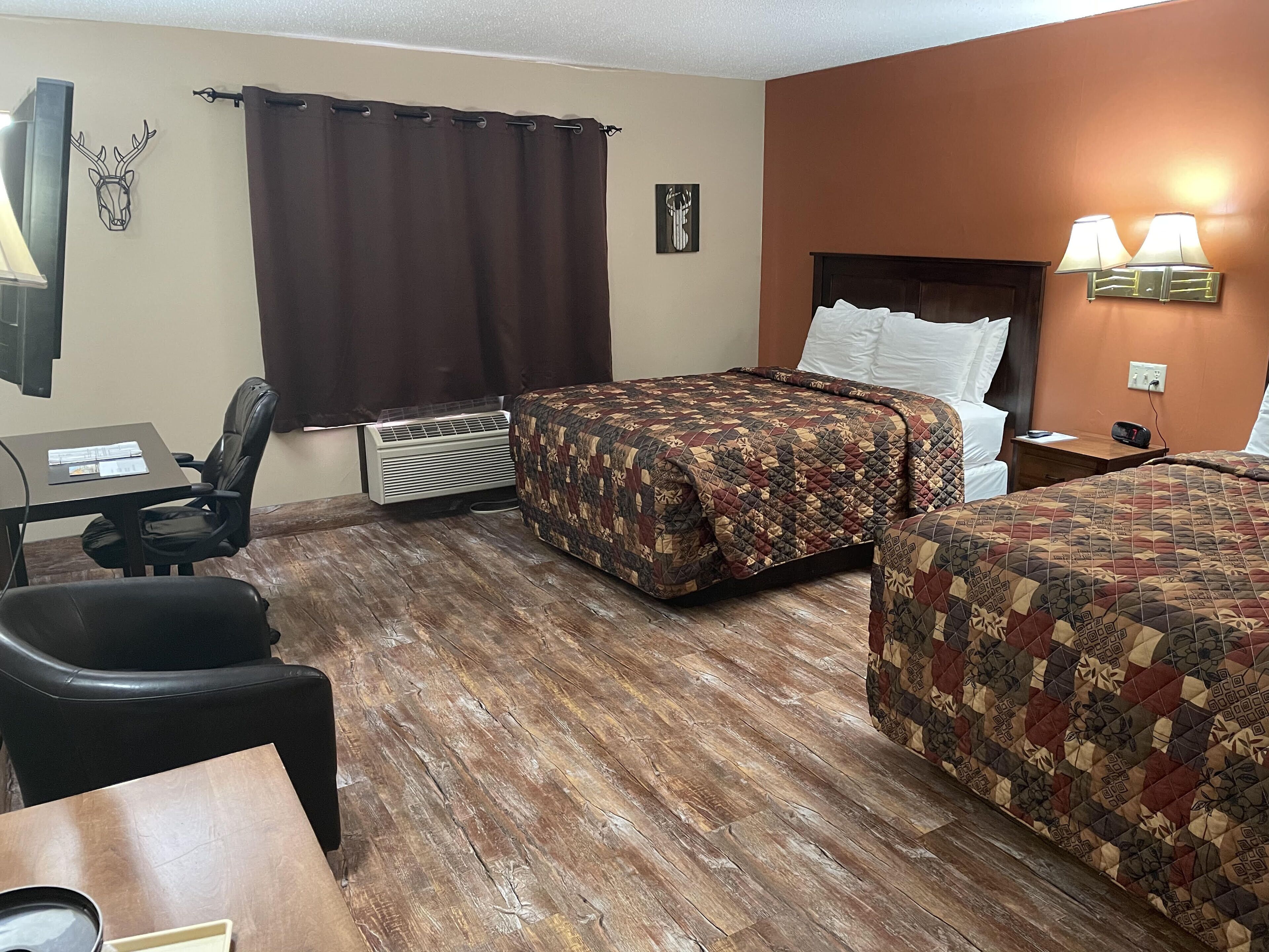 A hotel room with two beds, a desk, a chair, an air conditioner, and a wall-mounted lamp.
