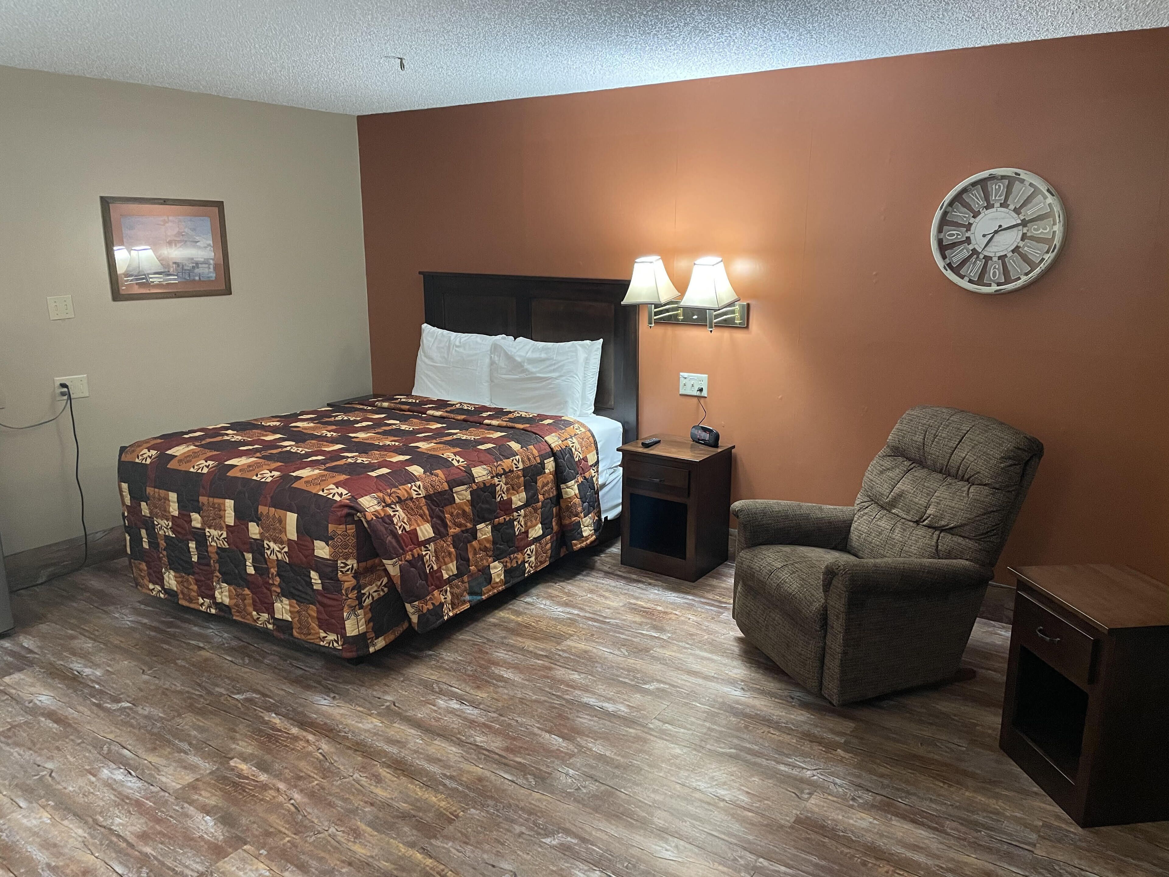Quarto de hotel com piso de madeira, cama com colcha estampada, uma cadeira, mesa de cabeceira com abajur e relógio de parede.