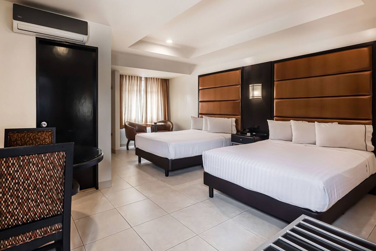 A hotel room with two beds, a seating area with chairs, a small table, and a wall-mounted air conditioning unit.