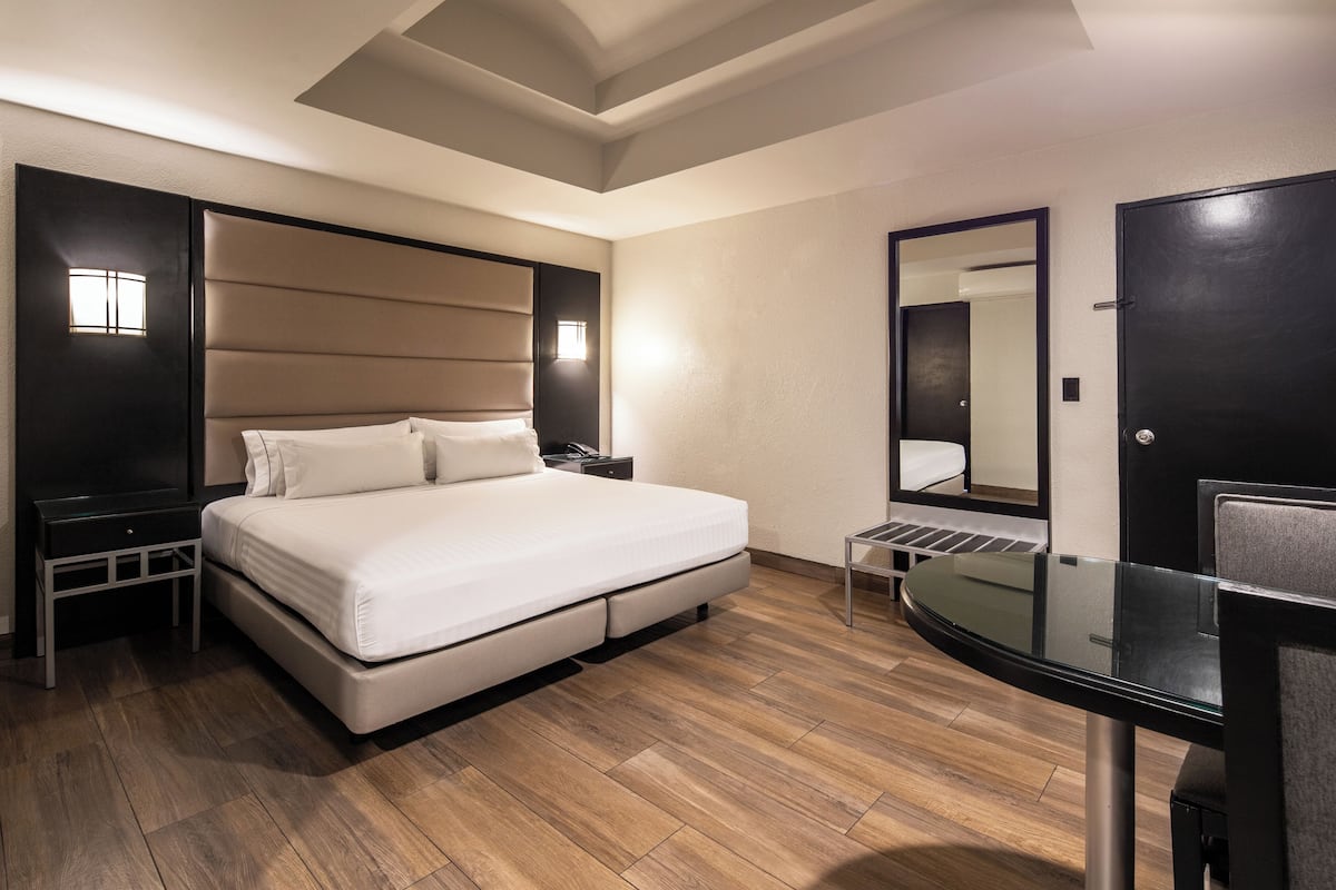 A modern hotel room with a large bed, a desk with a chair, a mirror, and a small table.