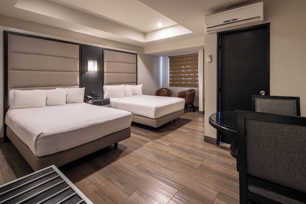 A hotel room with two beds, a desk, a chair, a window with blinds, and an air conditioning unit.