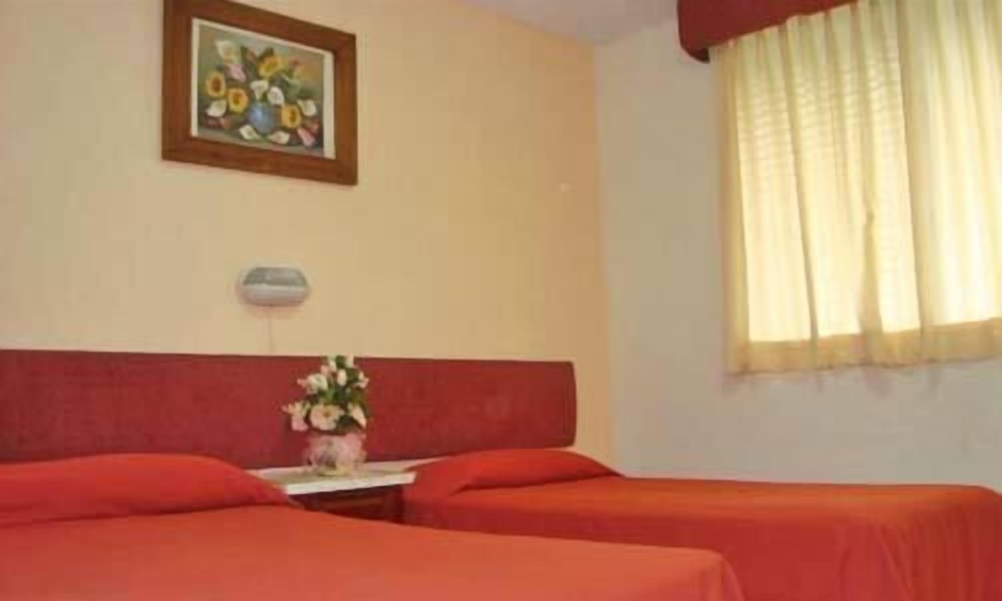 Quarto de hotel com duas camas, uma mesinha com um vaso de flores, um quadro na parede e uma janela com cortinas.