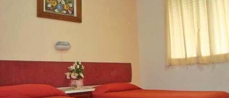 Quarto de hotel com duas camas, uma mesinha com um vaso de flores, um quadro na parede e uma janela com cortinas.