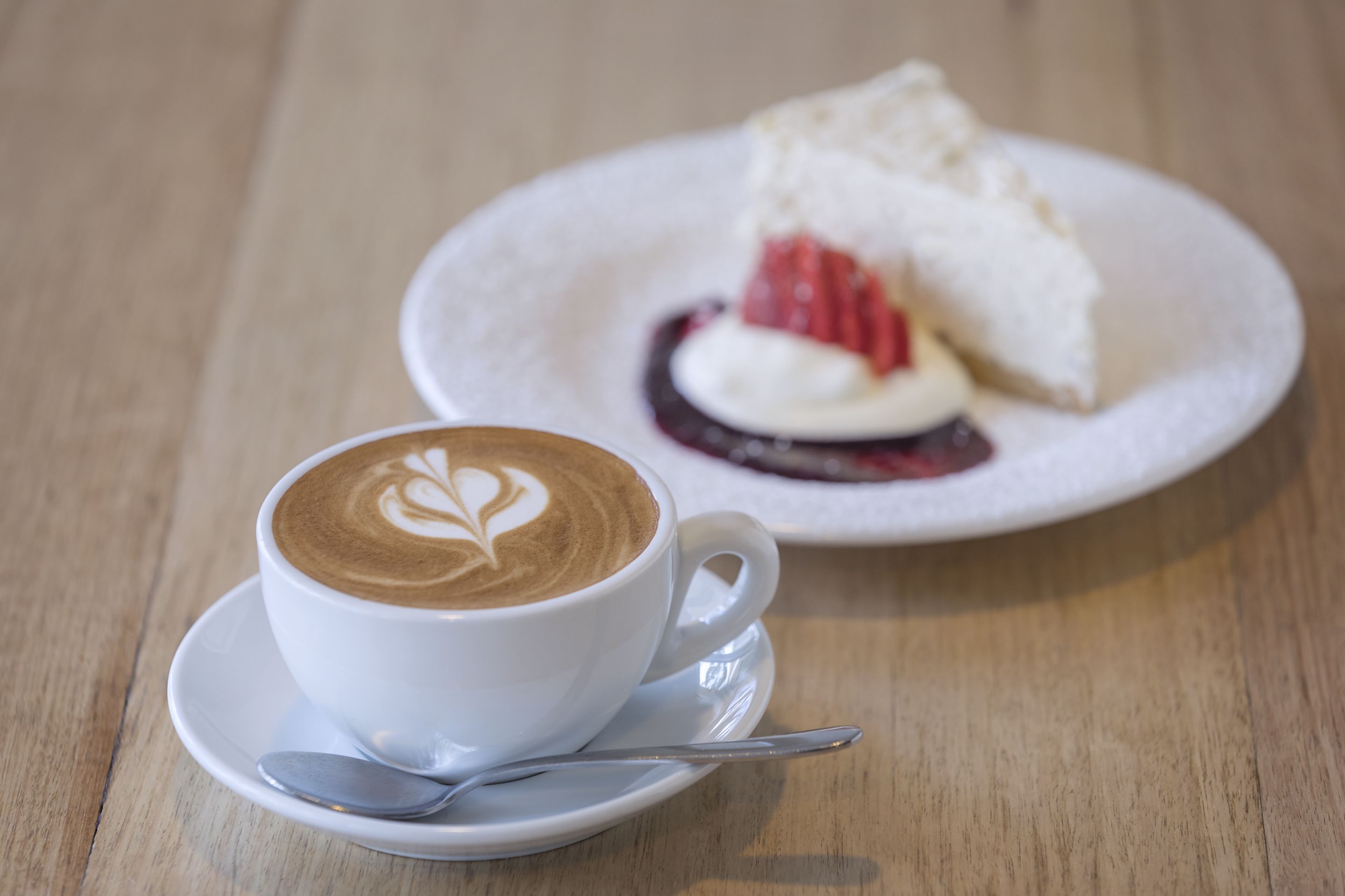 A white cup of coffee with latte art on a saucer, accompanied by a spoon, and a plate with a slice of cake and whipped cream.