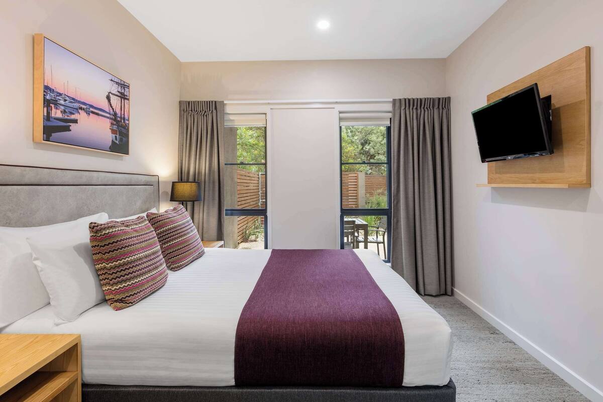 Ein Hotelzimmer mit Bett, einem an der Wand befestigten Fernseher, einem Gemälde über dem Bett und Blick ins Freie durch das Fenster.
