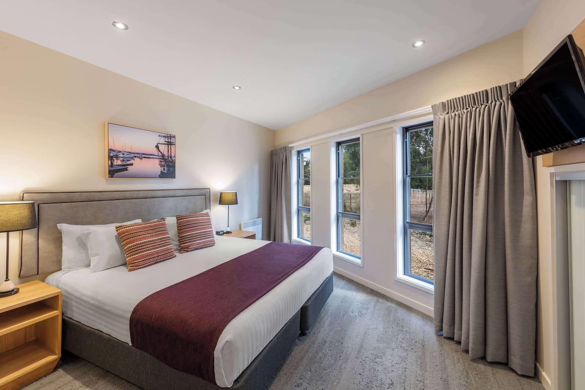 Ein Hotelzimmer mit einem großen Bett, einem flachbildfernseher an der Wand, einem Gemälde über dem Bett und einem Fenster mit Vorhängen.