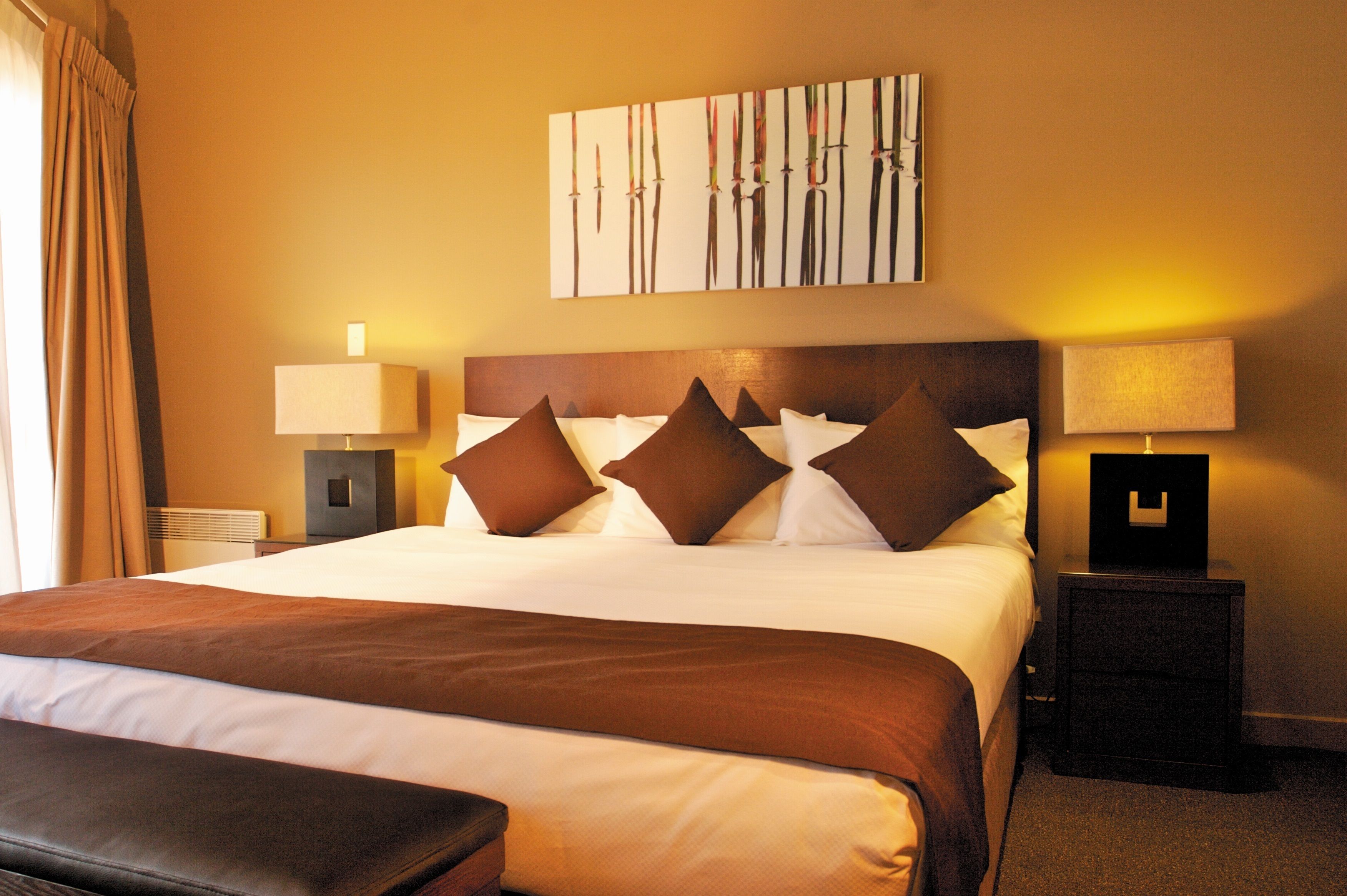 A hotel room with a large bed, a headboard with multiple pillows, a bedside table with a lamp, and a wall-mounted artwork.