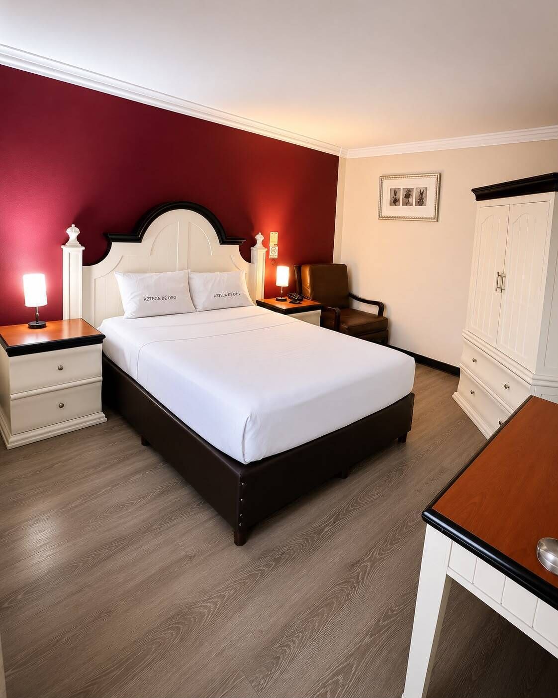 A hotel room with a bed, bedside tables, a desk, a chair, a wardrobe, and a nightstand with a lamp.