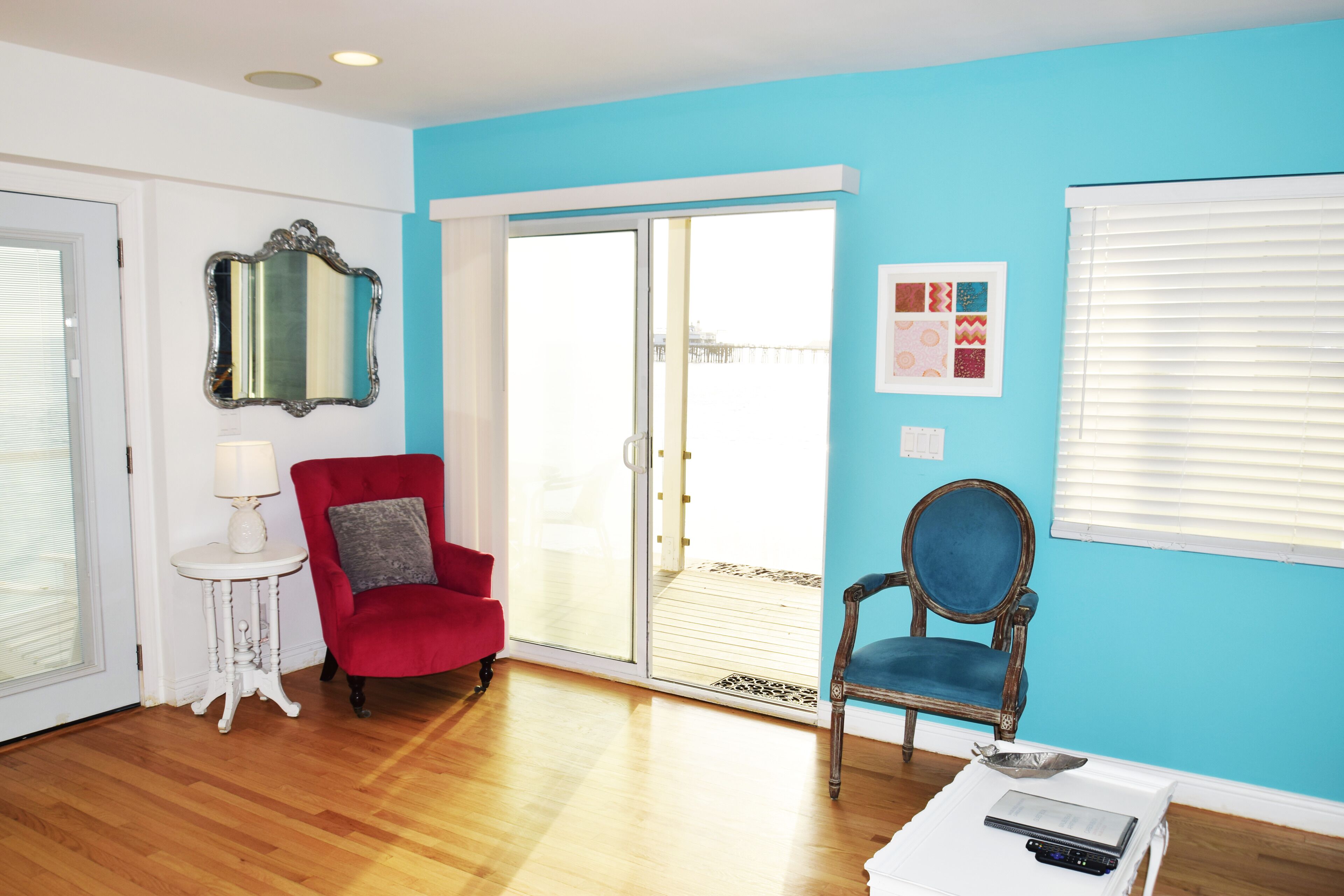 A room with a red armchair, a blue armchair, a white side table with a lamp, a mirror, and a sliding glass door leading to a balcony.