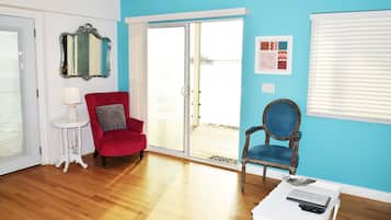 A room with a red armchair, a blue armchair, a white side table with a lamp, a mirror, and a sliding glass door leading to a balcony.