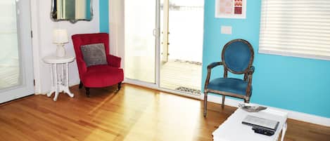 A room with a red armchair, a blue armchair, a white side table with a lamp, a mirror, and a sliding glass door leading to a balcony.