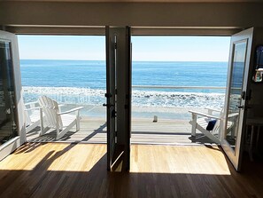 Ein Zimmer mit Holzboden, zwei weißen Stühlen und Blick auf das Meer durch geöffnete Türen.