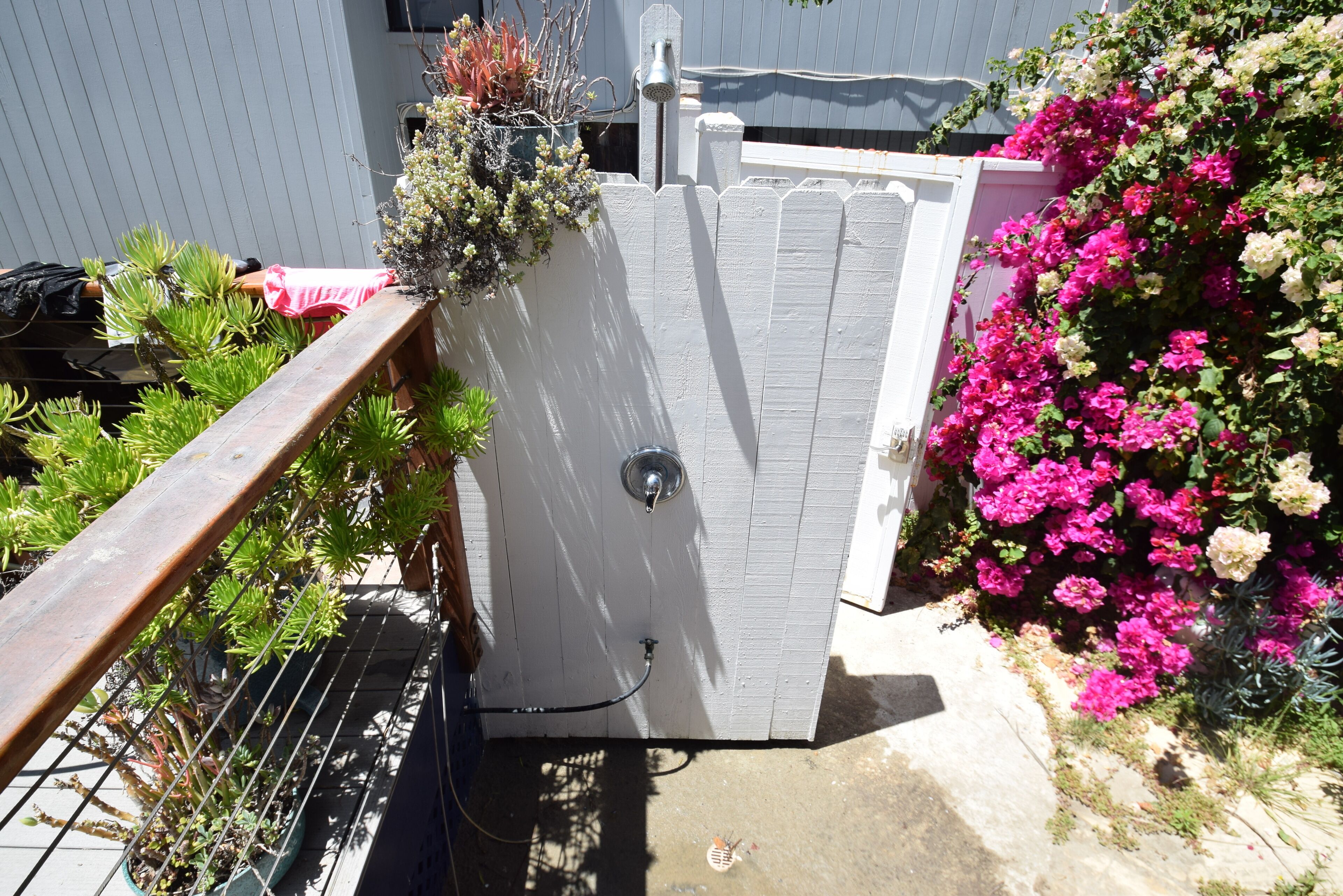 Un cobertizo blanco con puerta metálica, rodeado de exuberante follaje y plantas florecientes.