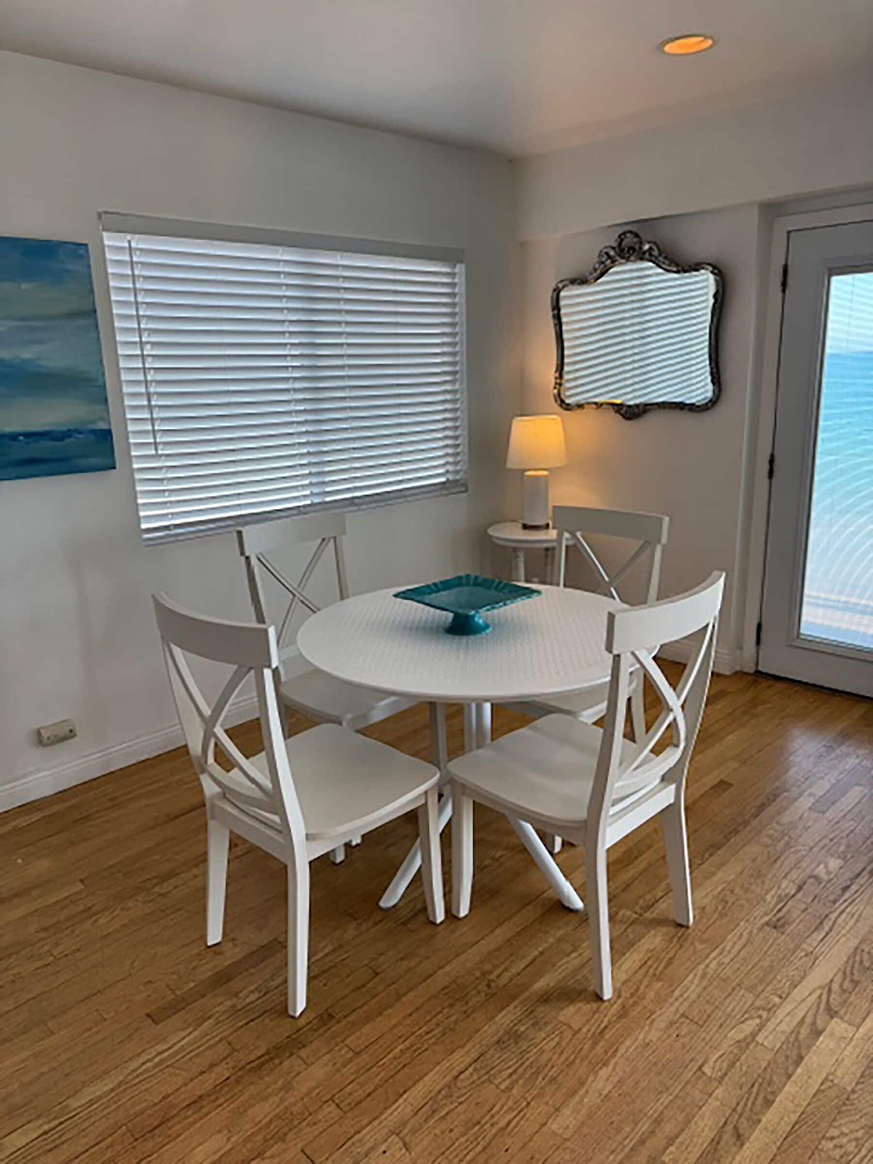 A dining area with a round table and four white chairs, a lamp on a side table, and a window with blinds.