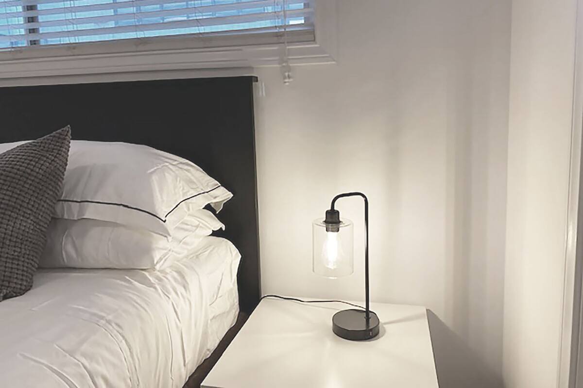 Een netjes opgemaakt bed met witte lakens en een grijs kussen, een nachtkastje met een zwarte lamp, en een raam met blinds.
