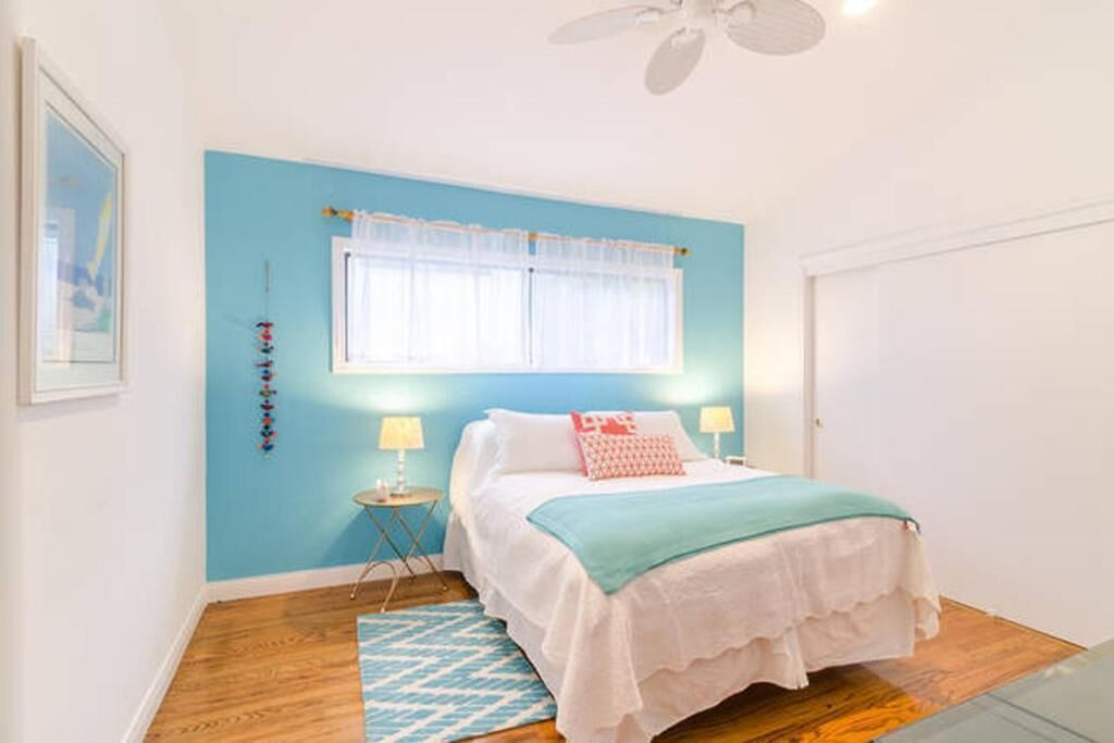 Un dormitorio con cama, ventana con cortinas, ventilador de techo y una mesita de noche con lámpara.
