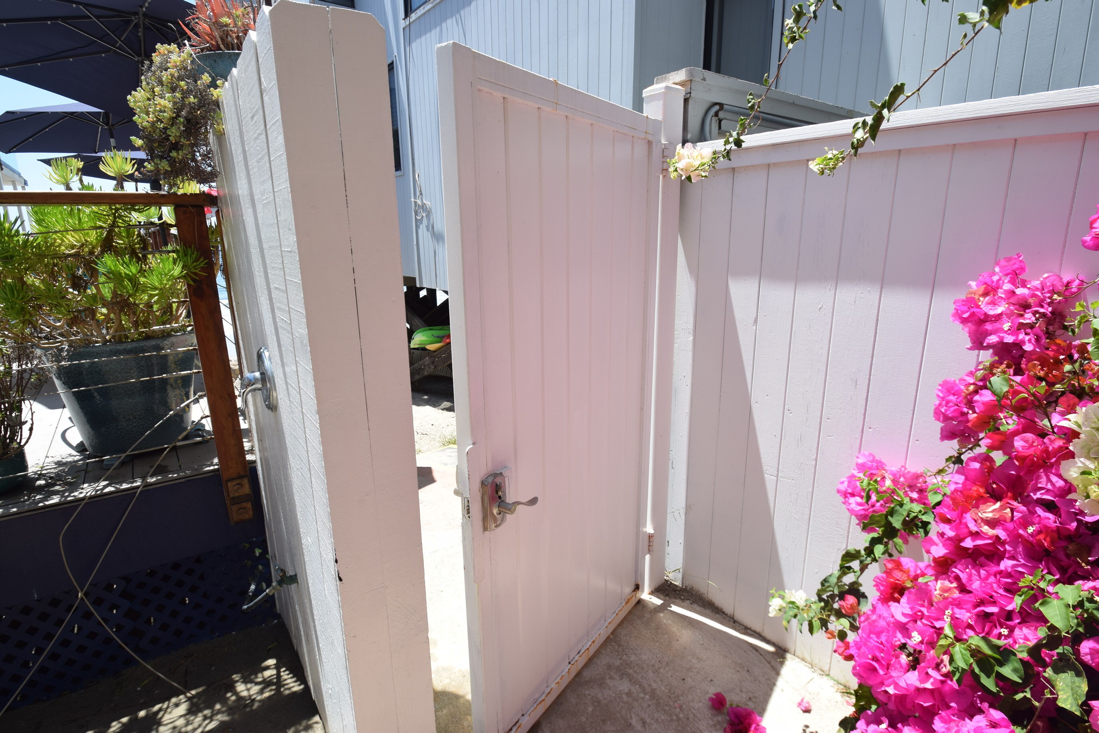 A white wooden gate with a metal latch, leading to a covered patio area with potted plants and a concrete floor.