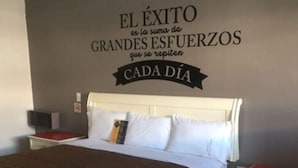 Een slaapkamer met een bed, witte nachtkastjes, een rode accentkleur op de nachtkastjes, een muur met een motiverende quote, en een bruine sprei.