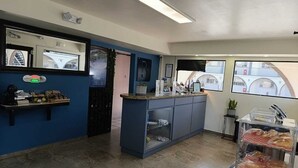 Een hotel receptie met een blauwe muur, een receptiebalie, een klein servicegedeelte, een raam met uitzicht op gebouwen en een presentatie van gebak.