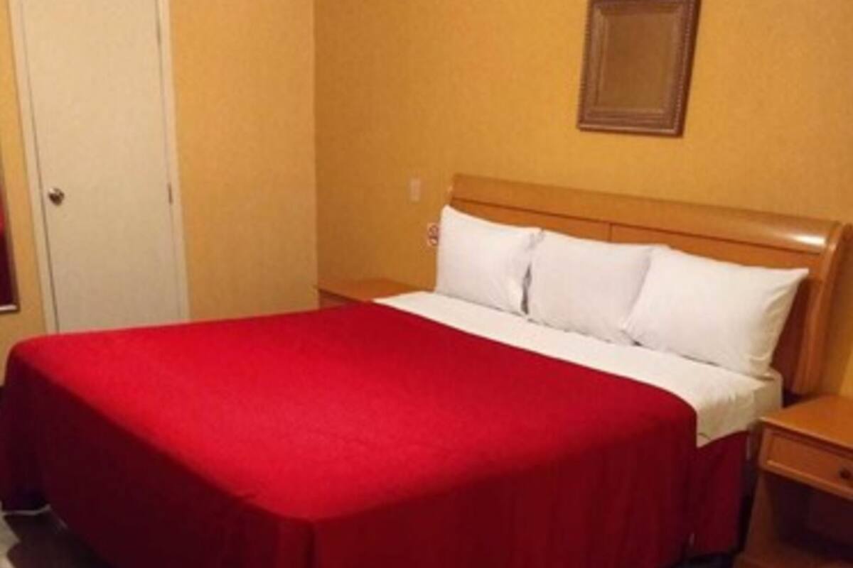 Quarto de hotel padrão com estrutura de cama de madeira, roupa de cama branca e colcha vermelha.