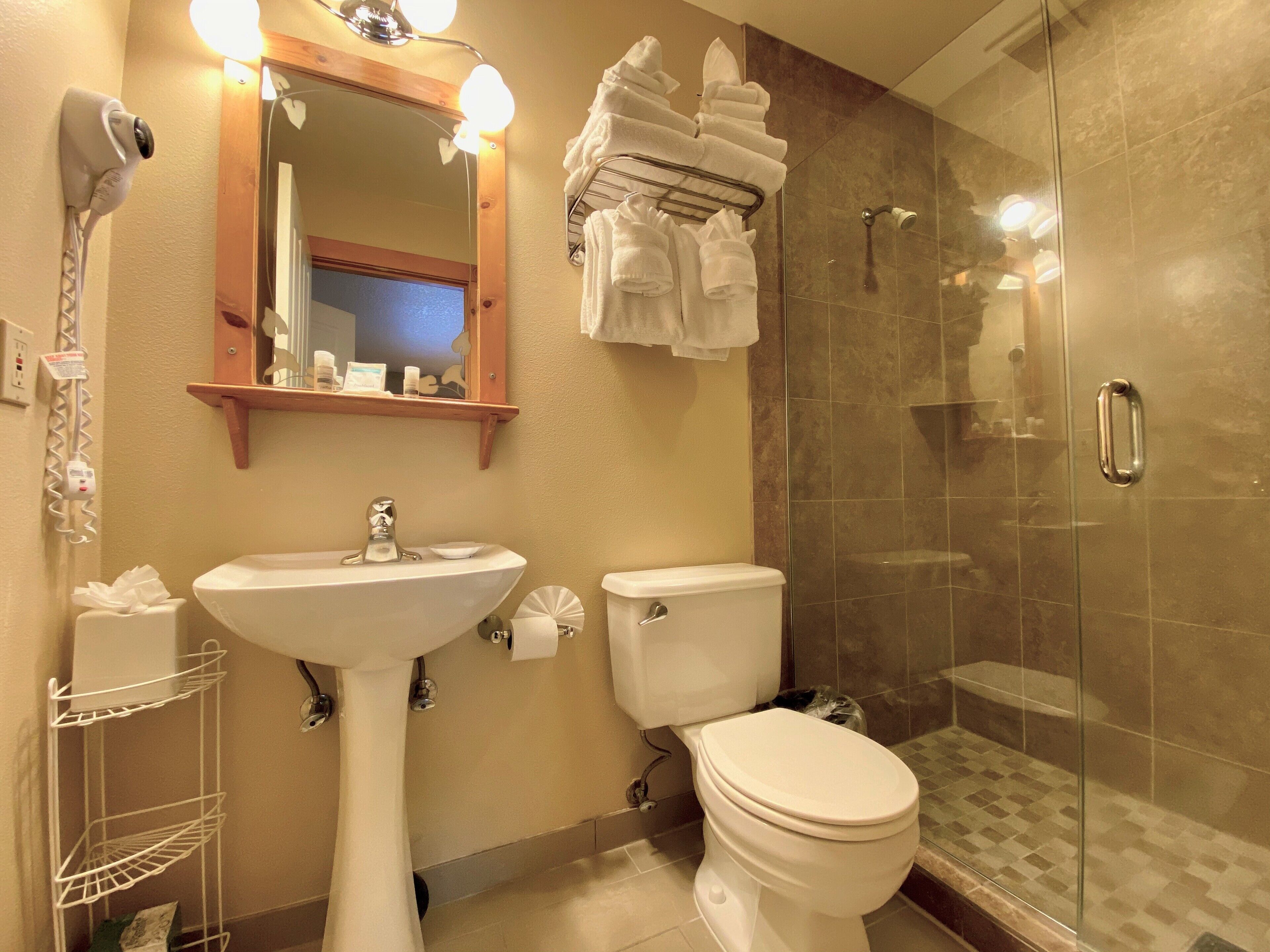 A bathroom with a pedestal sink, a toilet, a glass shower enclosure, a towel rack with folded towels, a mirror with lights, and a hairdryer on the wall.