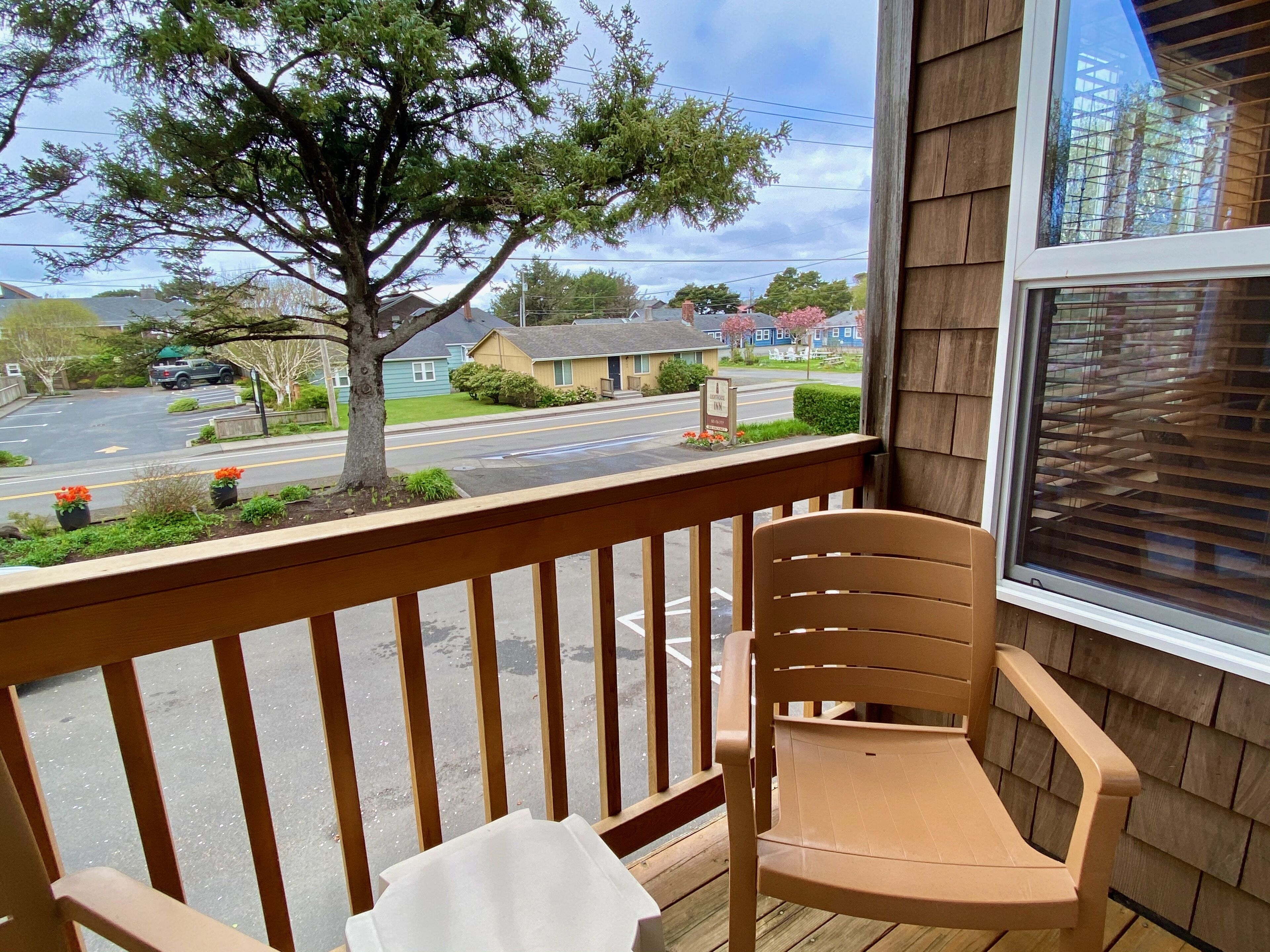 A wooden deck with a single chair and a table, overlooking a street with houses and a tree.