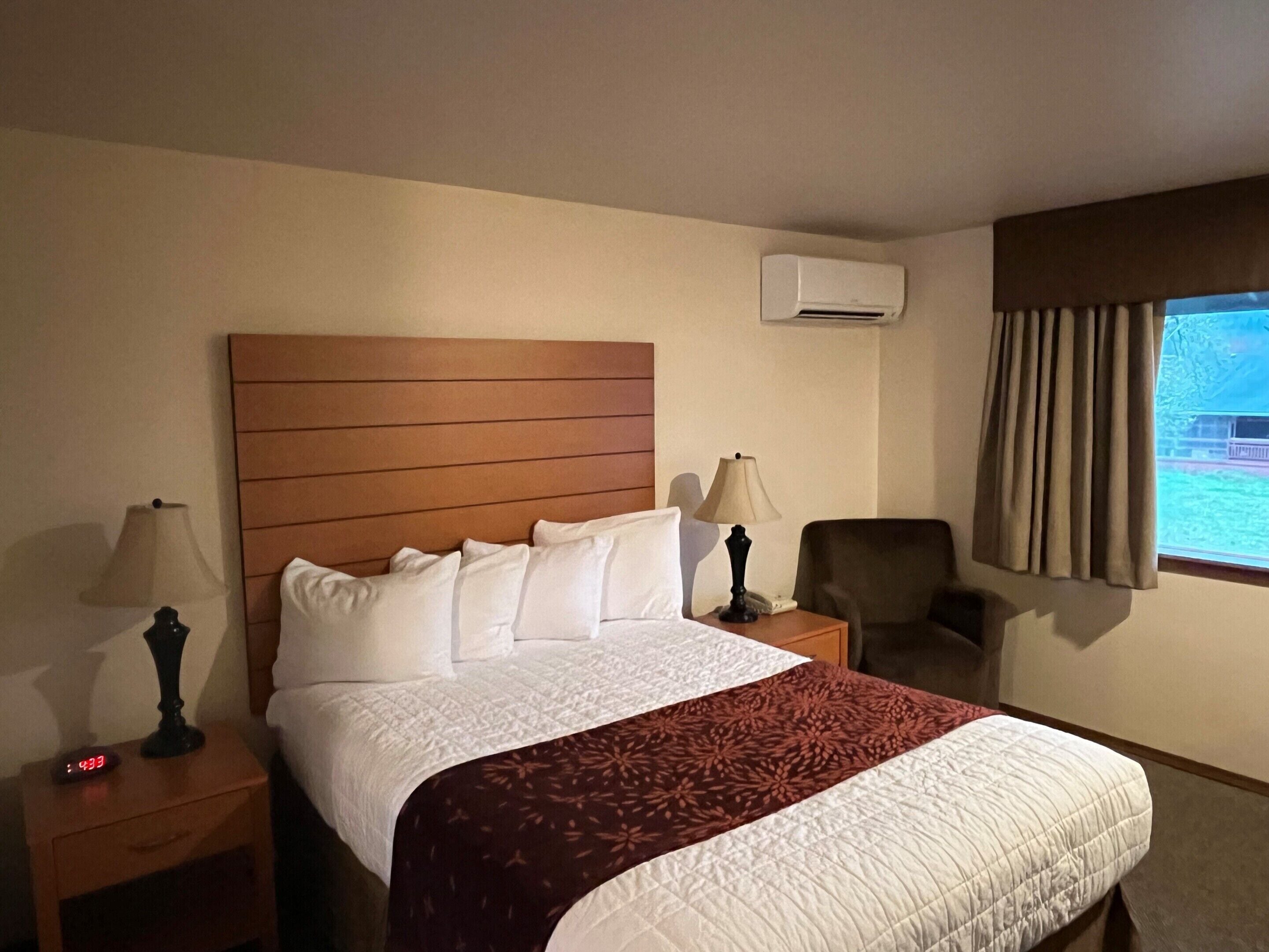 Habitación de hotel con cabecera de madera, cama con ropa de cama blanca y granate, dos lámparas de noche, una silla, una ventana con cortinas y un equipo de aire acondicionado.