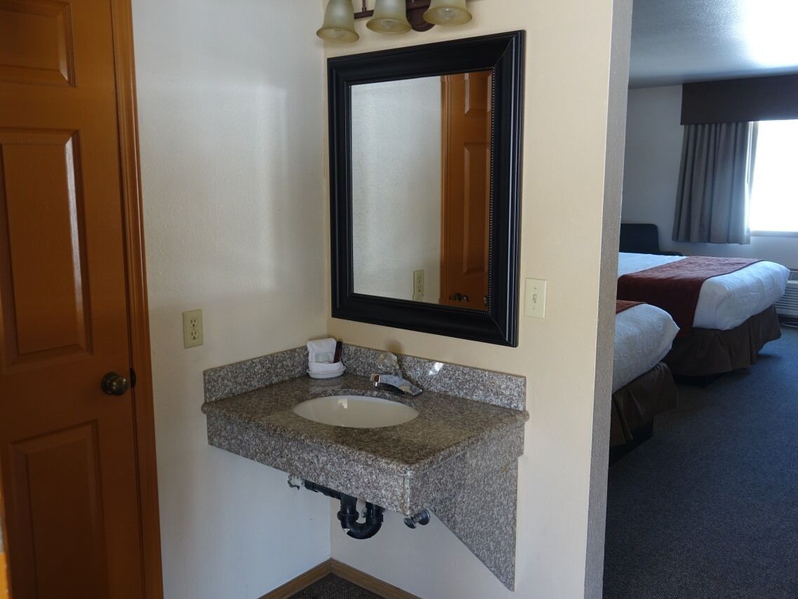 Un baño de hotel con lavamanos de encimera de granito, un espejo y una puerta que da a un dormitorio con dos camas.