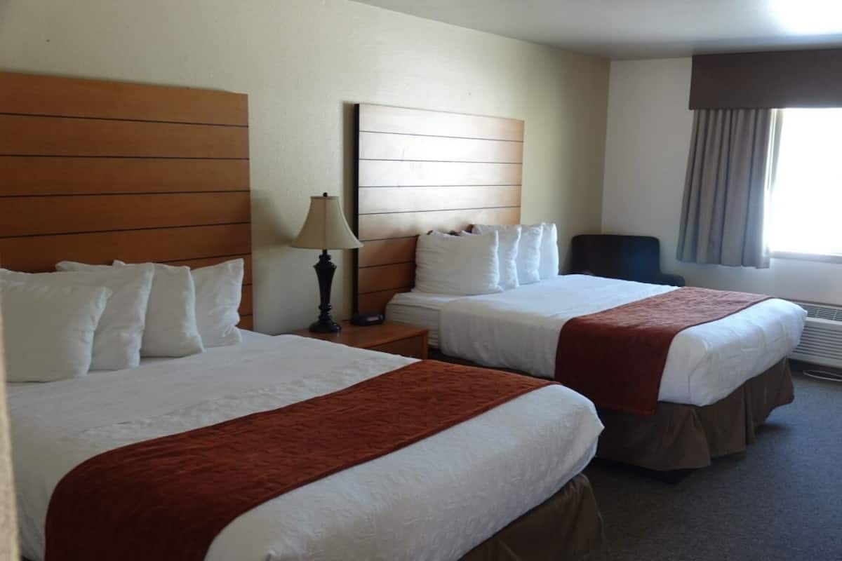 A hotel room with two beds, a nightstand, a lamp, a chair, and a window with curtains.