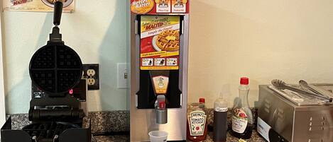 A self-serve waffle station with instructions, condiments, and a waffle maker on a kitchen counter.