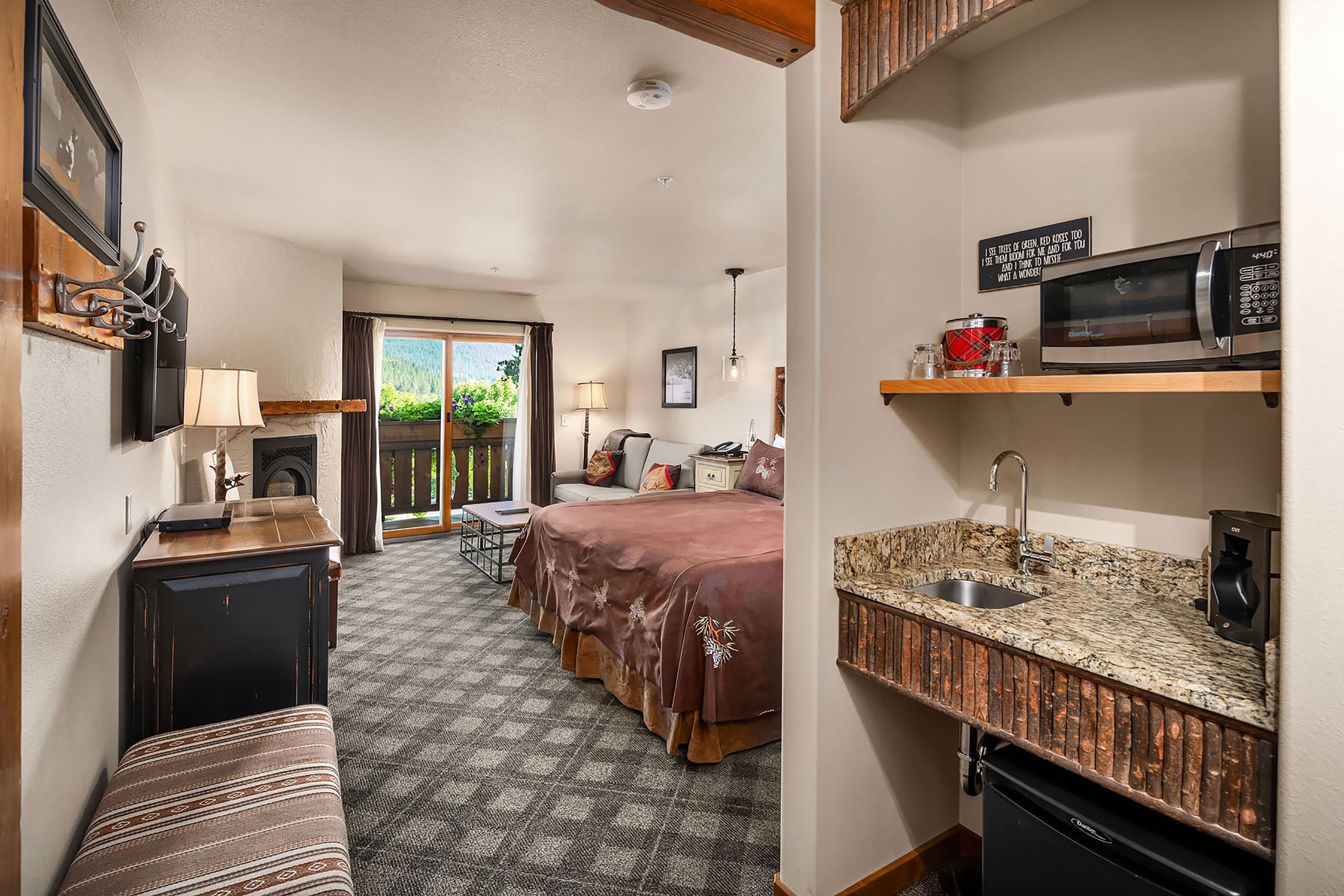 A hotel room with a bed, a desk, a chair, a microwave, a sink, and a window with a view of greenery.