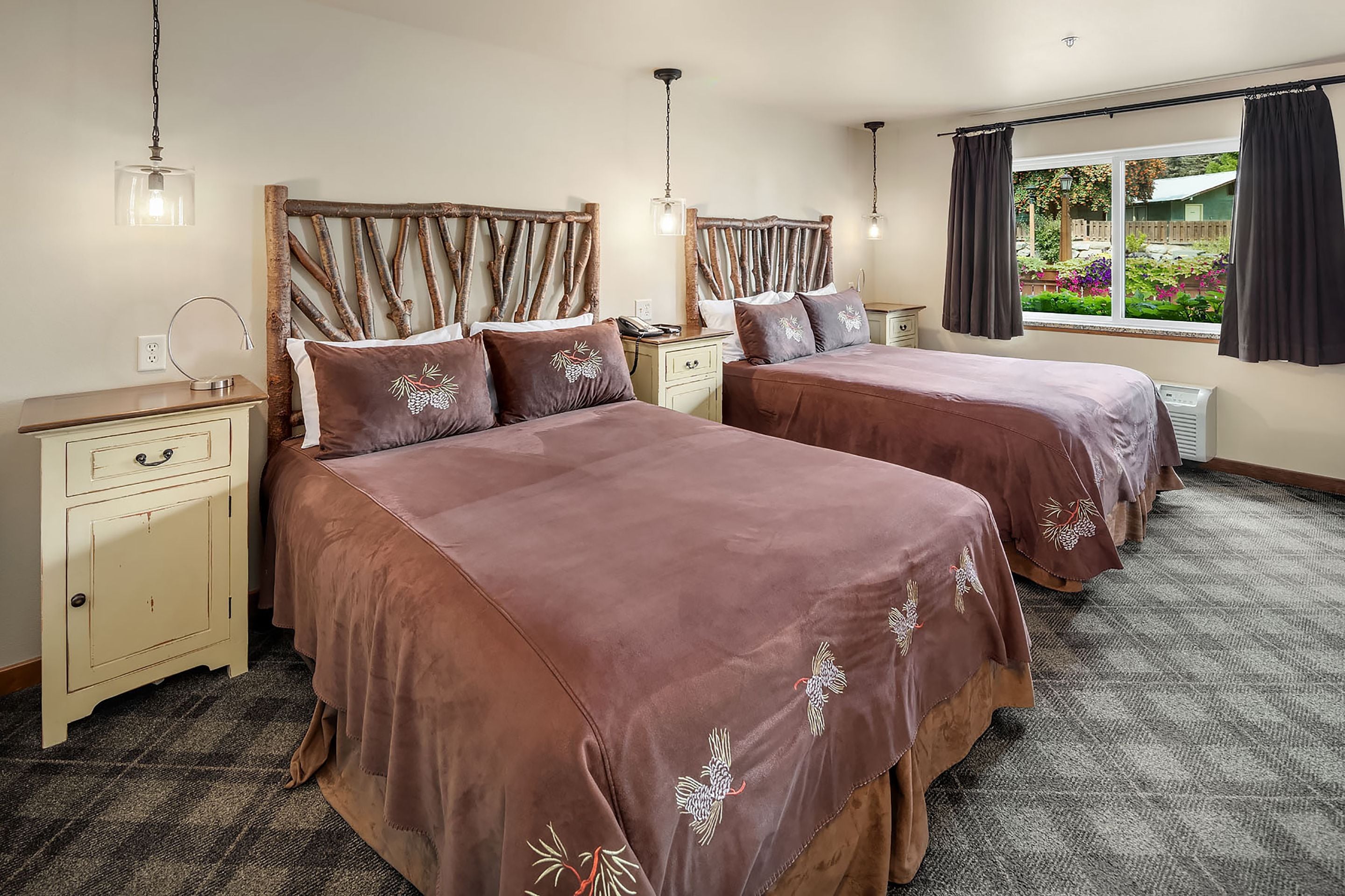 A hotel room with two beds, a nightstand, a lamp, a window with curtains, and a view of a garden.