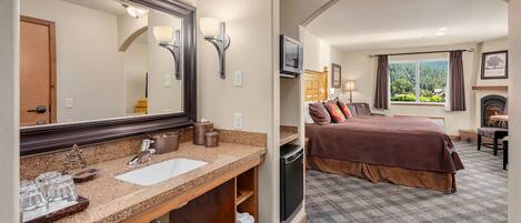 A hotel room with a large bathroom vanity, a bed with a brown bedspread, a television, and a fireplace.