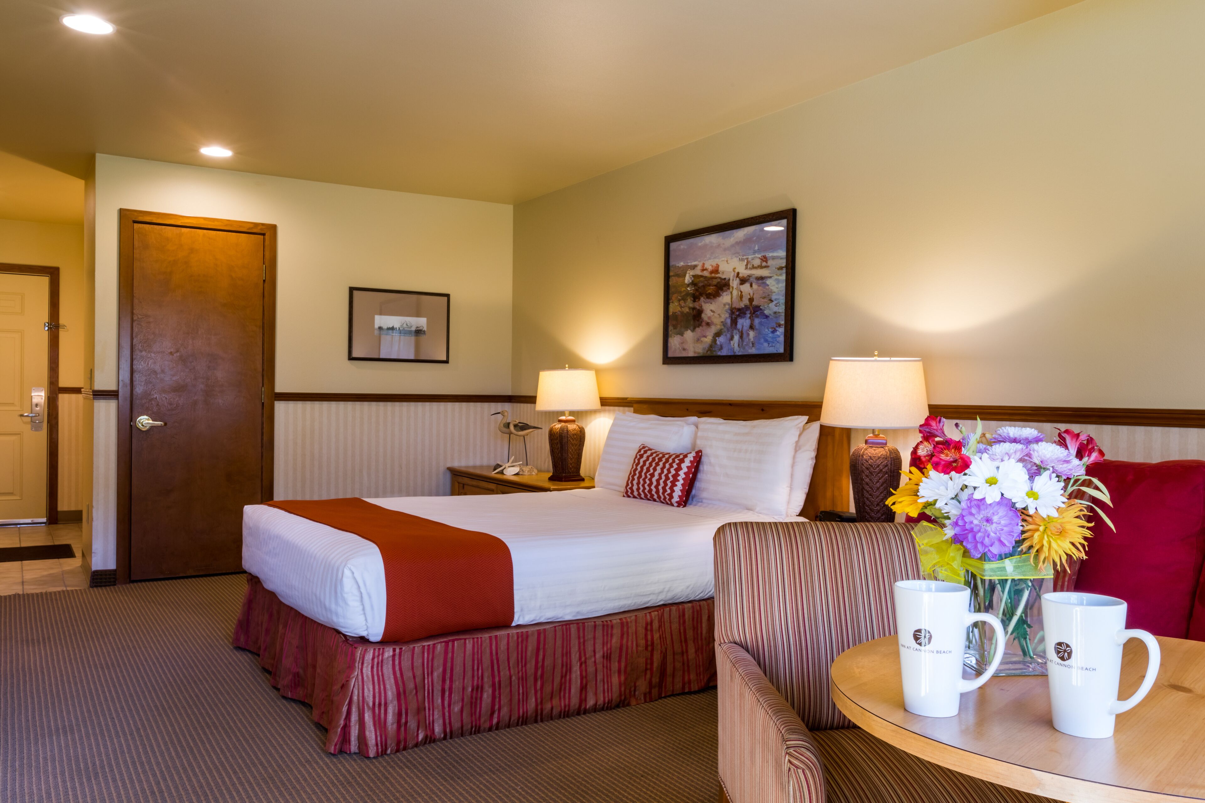 A hotel room with a bed, a chair, a table with two mugs, a lamp, a framed picture, and a door.