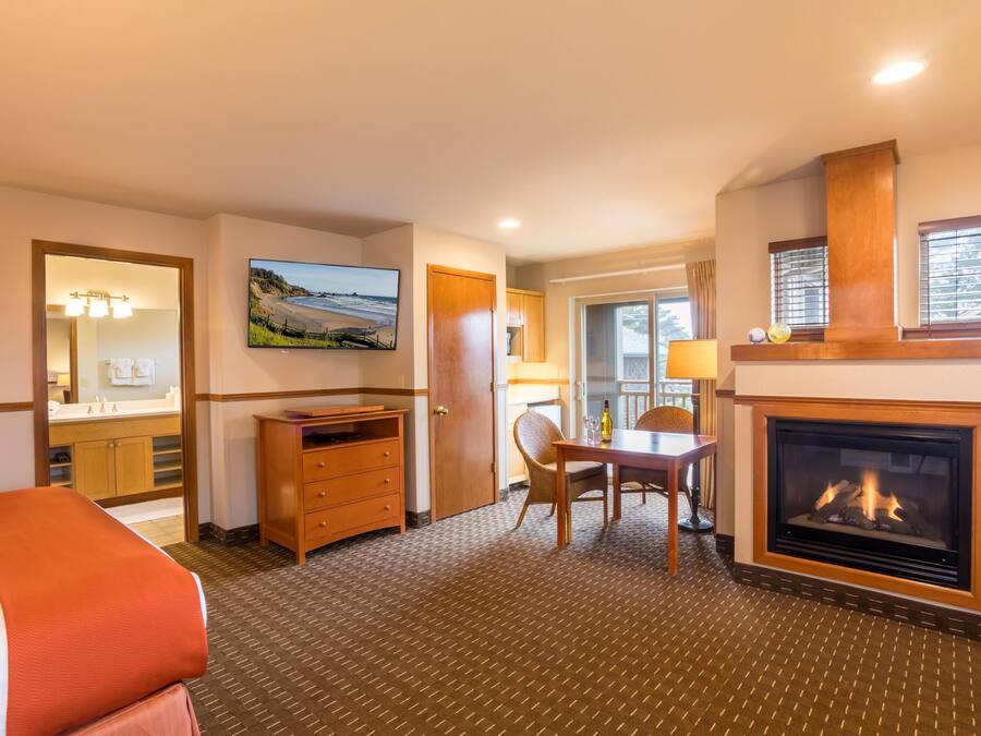 A hotel room with a bed, a fireplace, a dining table with chairs, a TV, and a bathroom visible through an open door.