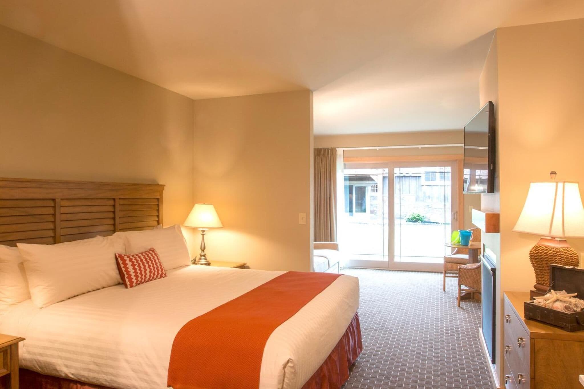 A hotel room with a large bed, a television, a desk with a lamp, and a view of the outside through a glass door.
