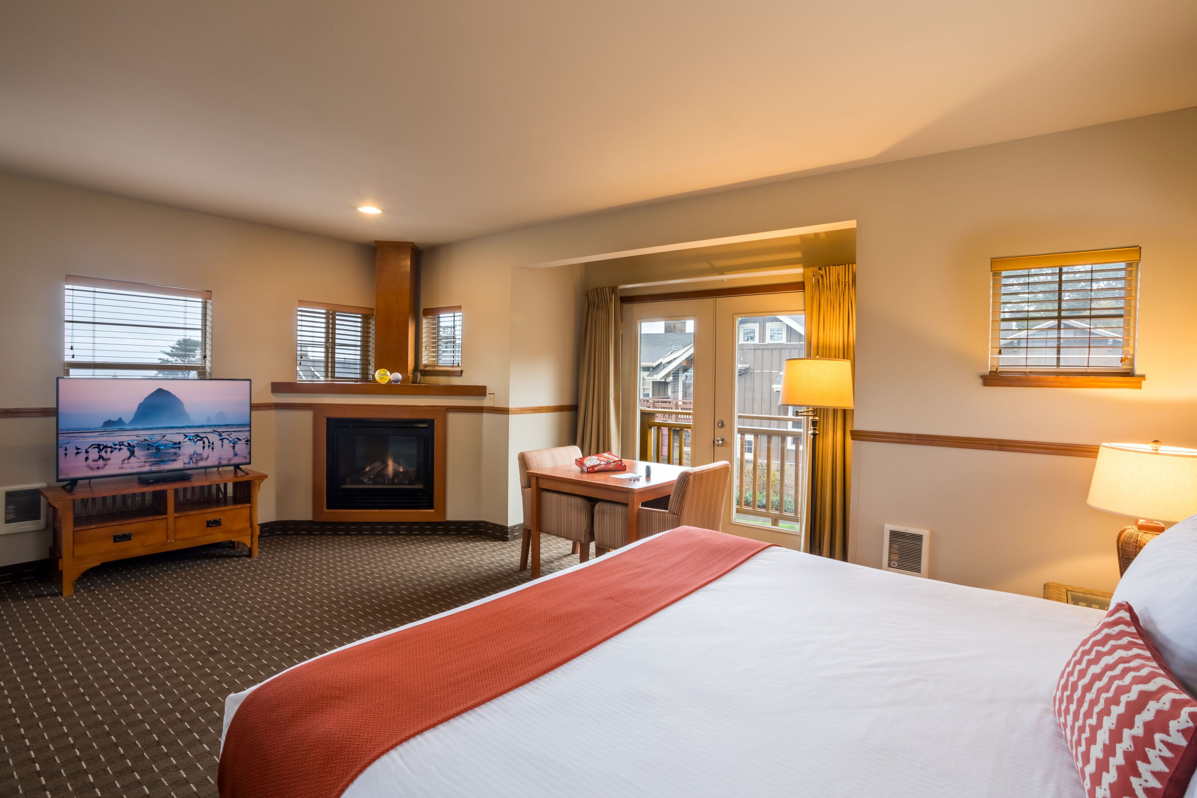 A hotel room with a large bed, a TV on a wooden stand, a fireplace, a balcony with a view, and a desk with a chair.