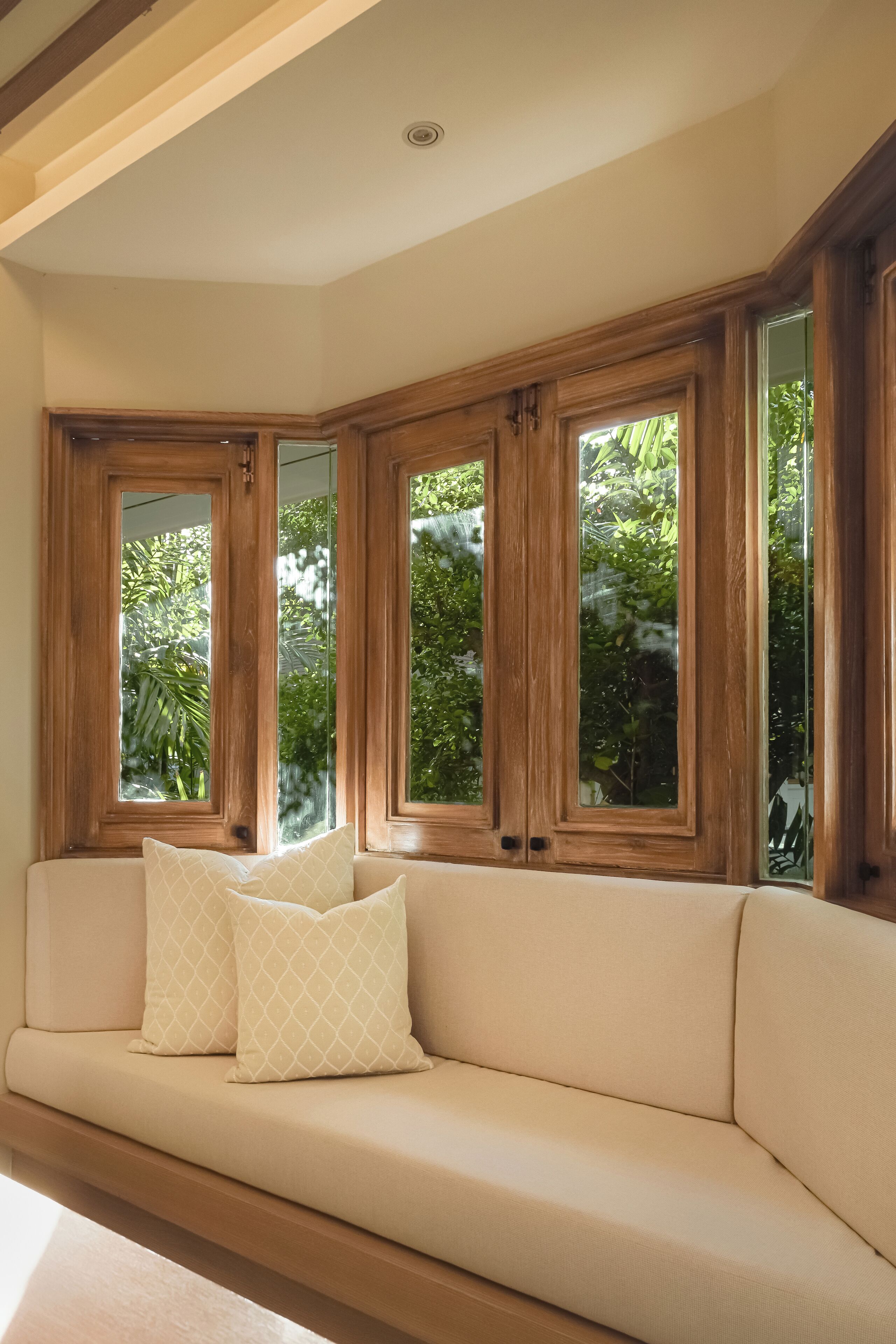 Un sofá color beige con dos cojines estampados, ubicado debajo de ventanas con marco de madera y vistas a zonas verdes.