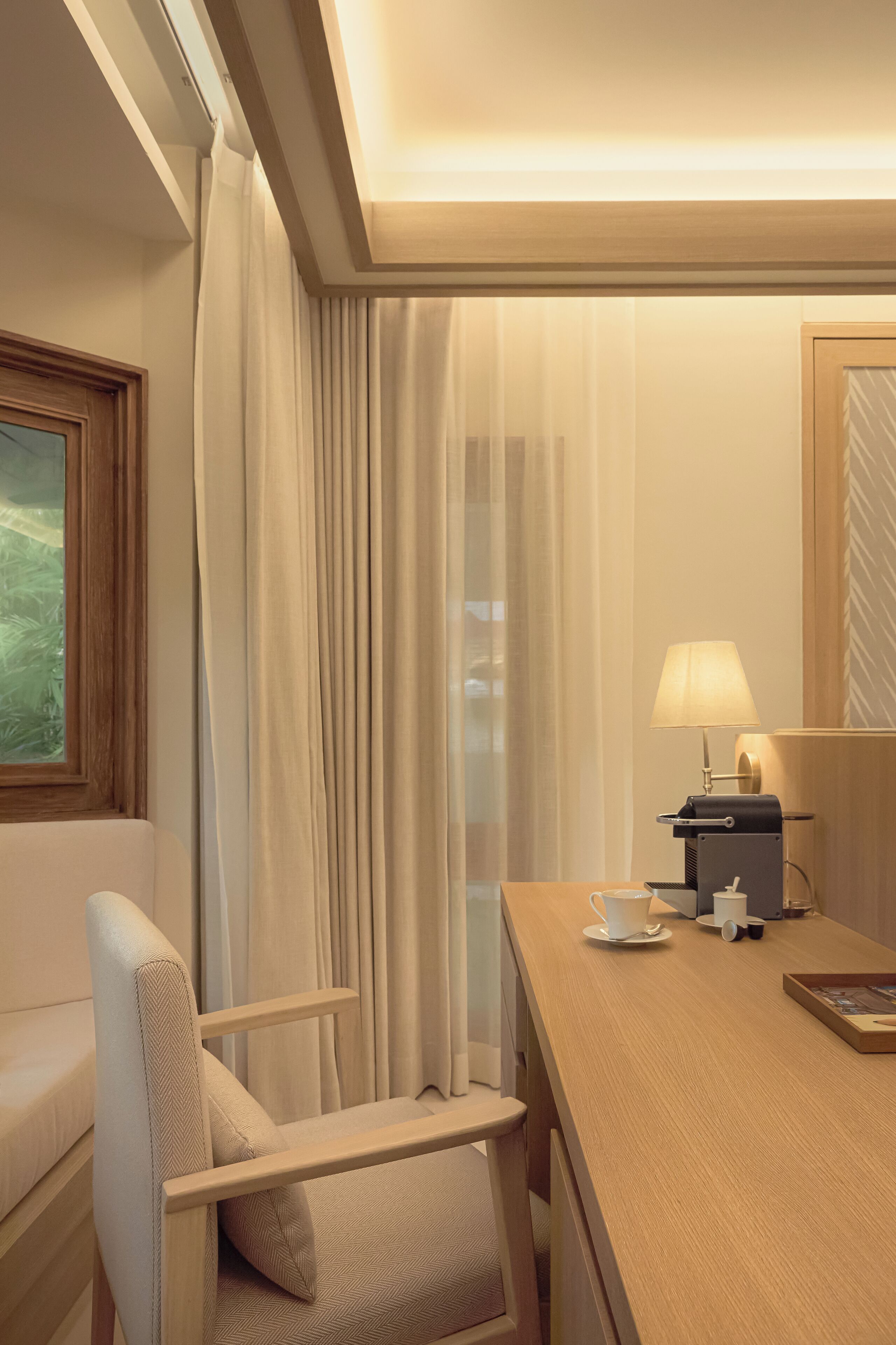 A hotel room with a wooden desk, a lamp, a coffee maker, and a view of greenery through the window.