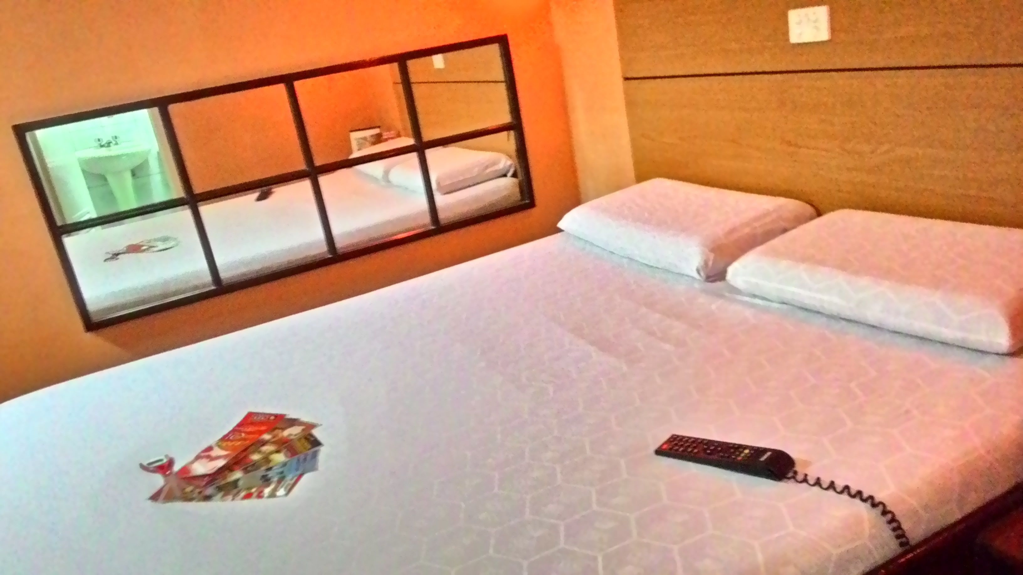 Una habitación de hotel con una cama, un ventanal con vistas a un lavamanos y un espejo, y un control remoto sobre la cama.