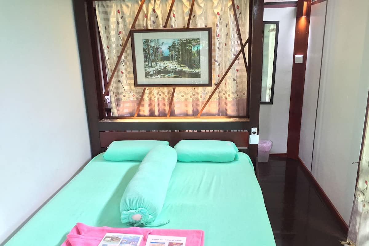A neatly made bed with green bedding and a pink towel, a framed picture on the wall, and a window with curtains.