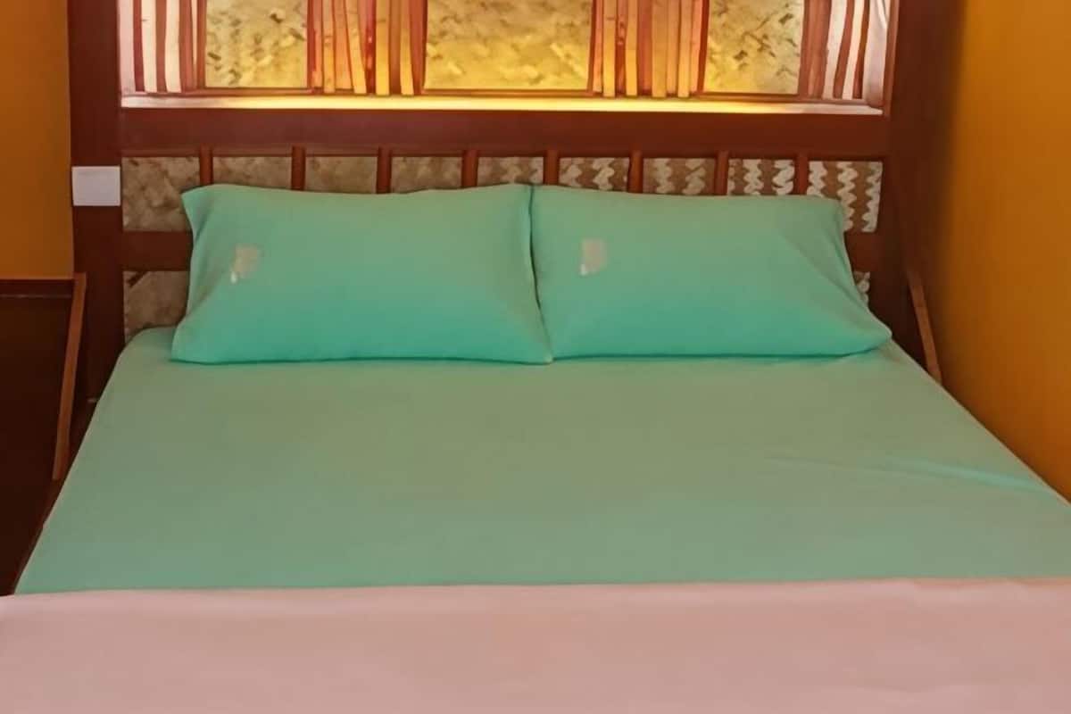 A bed with a green bedspread and two pillows, a wooden headboard, and a window with a patterned curtain.