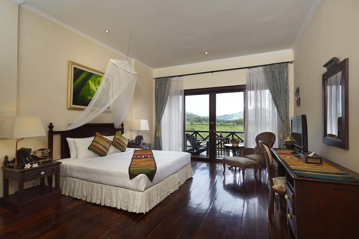 A spacious bedroom with a large bed, a nightstand, a desk, and a balcony with a view of the hills.